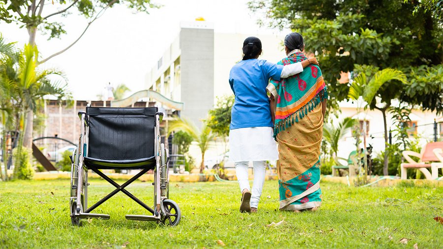 A healthcare worker supports an elderly woman as they walk together in a garden, with an empty wheelchair left behind on the grass.