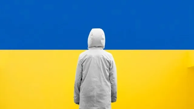 A person in a hooded jacket stands facing the Ukranian flag. 