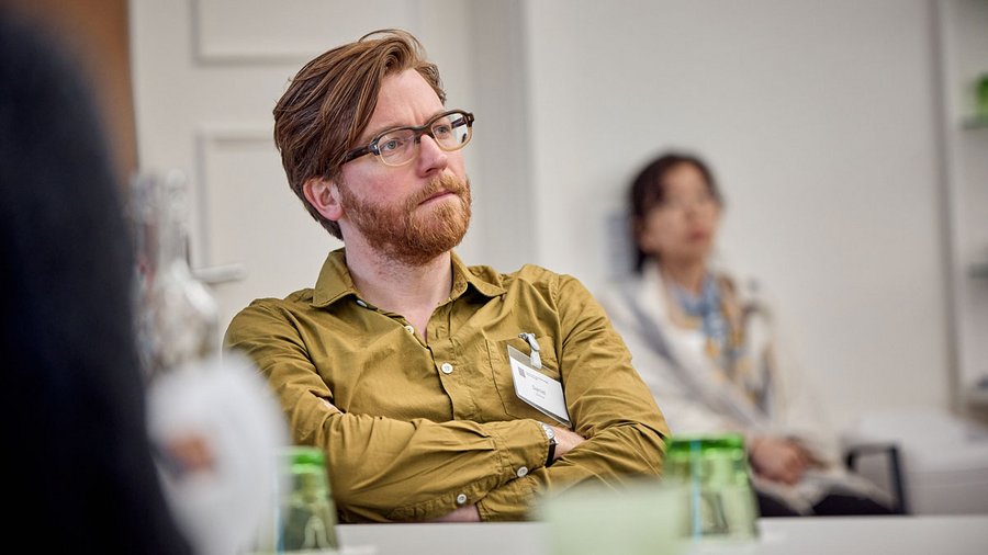 Daniel Gorman, a man wearing a brown shirt and glasses, sits at a table with his arms crossed