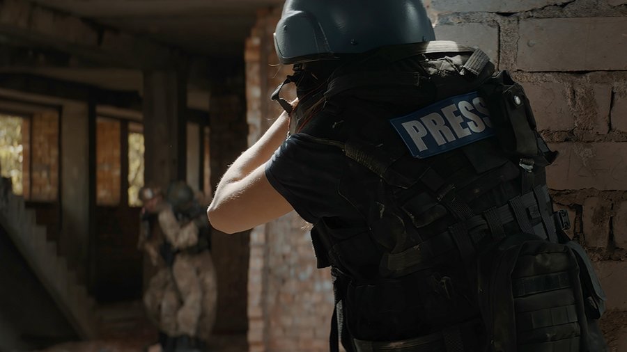 A journalist wearing a vest labelled "press" peers around a brick corner at soldiers