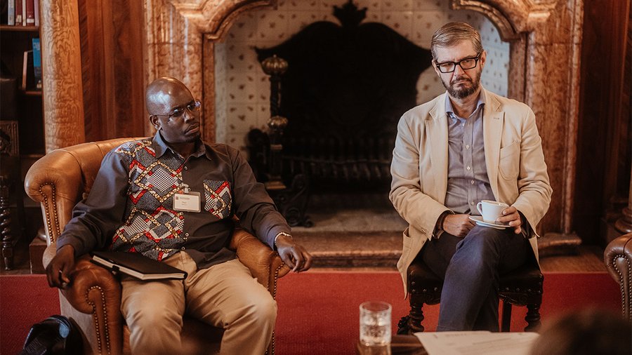 This image shows João Marcos Paes Leme sitting to the right of another man and looking attentively at another speaker who is off-camera.