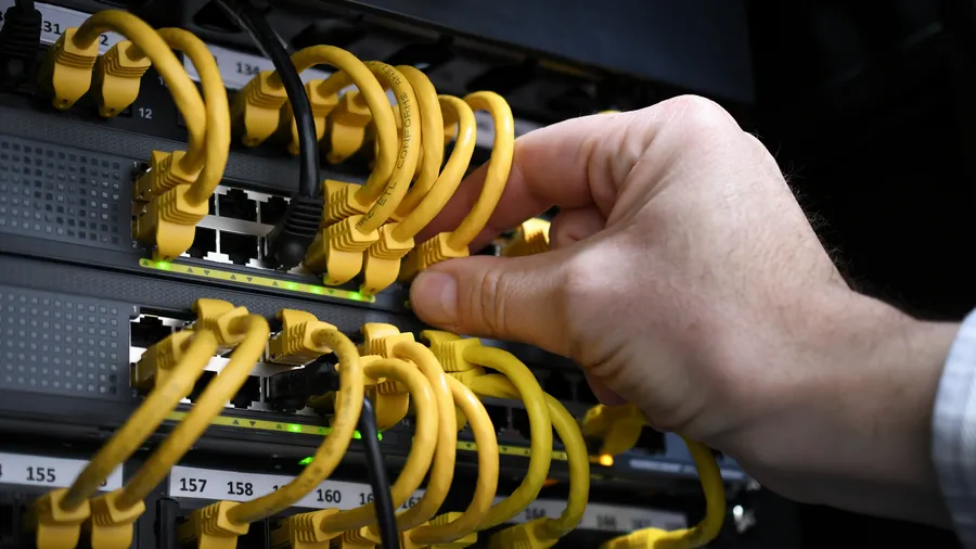 A hand reaches into a server room to connect a yellow cable into the network. The close up shot highlights the high tech nature of the server room and the complex network of cables and equipment.