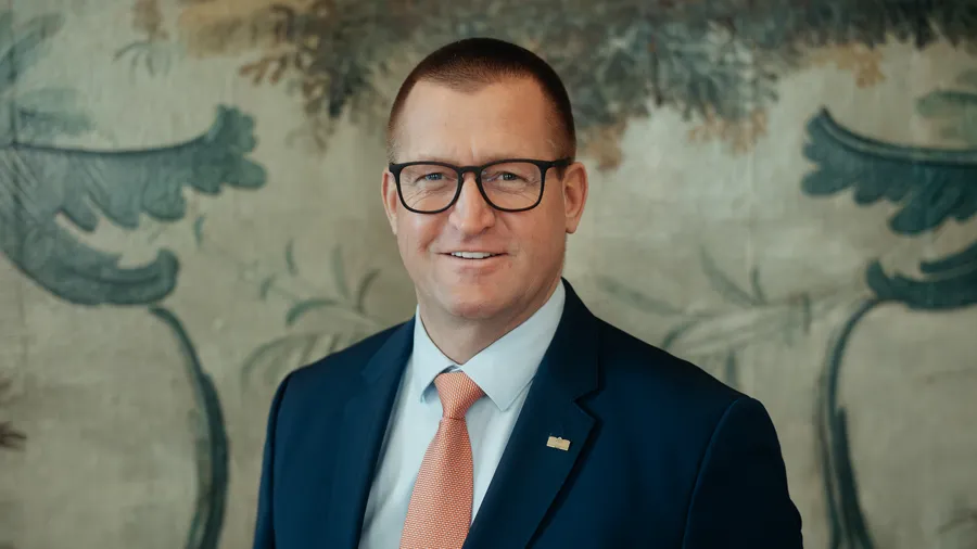 The image depicts a well-dressed individual standing against a backdrop of an ornate, floral patterned wall.