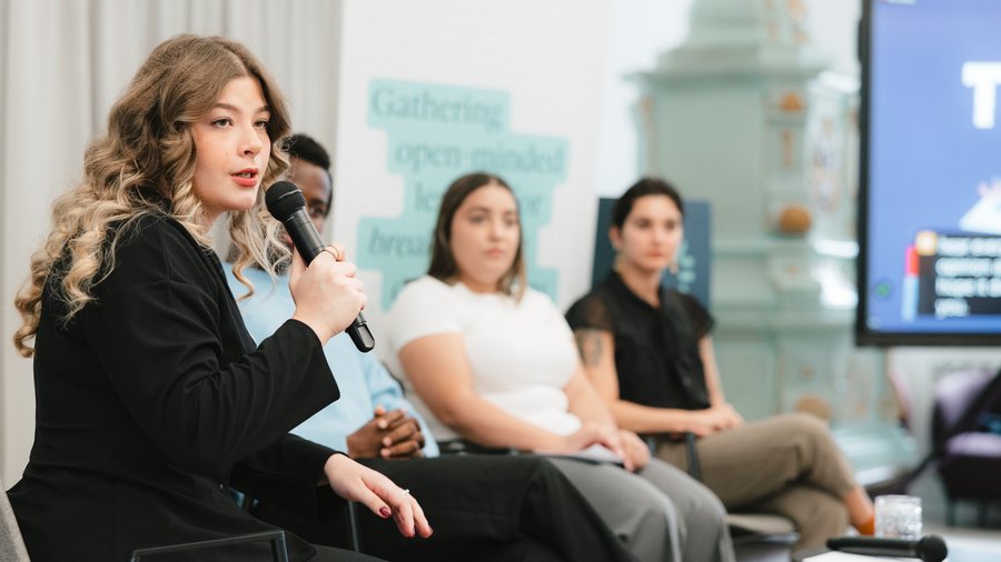 A woman with curly hair speaks into a microphone with other people seen in the background.