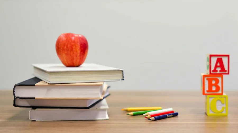 An apple sits atop a stack of books on a desk, with a few crayons and alphabet blocks on its right side.
