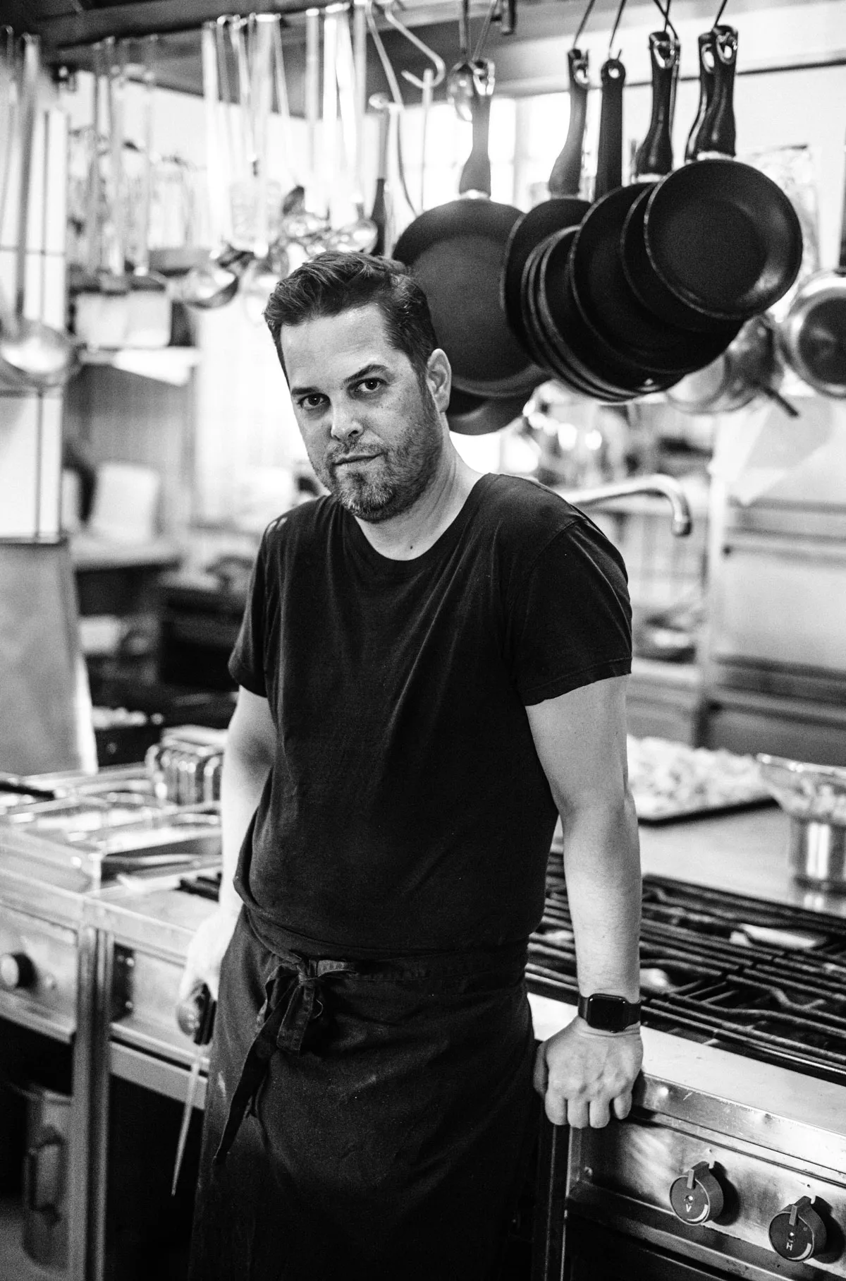 Black-And-White Photo of a Man in a Kitchen With Pans Hanging From the Ceiling in the Background