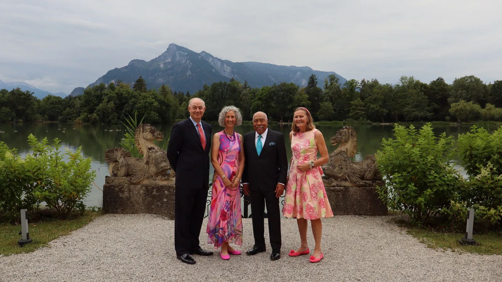 four people, two men and two women, dressed in black tie stand together in front of a lake with mountain scenery in the back.