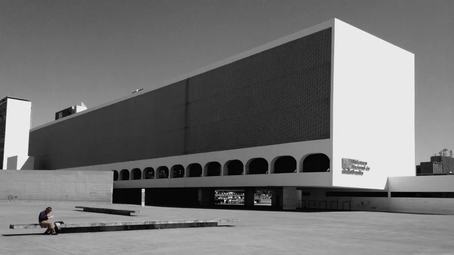 The image shows a man sitting with his lunch box eating his lunch, in the hot sun, in contrast with the buildings behind, which is also a public library.