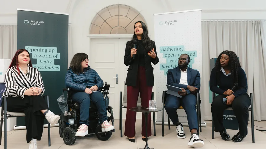 a woman with dark hair speaks into a microphone while standing next to four other individuals