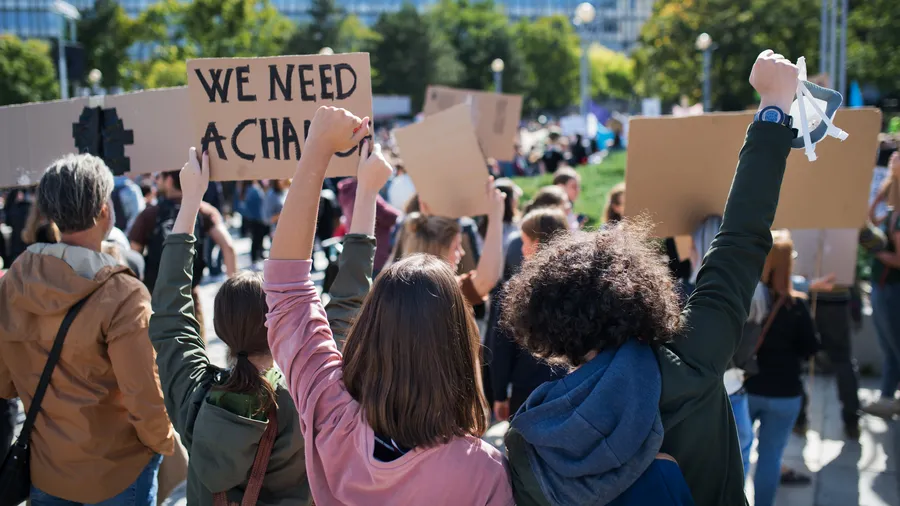 A crowd of people are gathered outdoors in the city on a sunny day. They are holding signs and raising their arms, appearing to be part of a protest or rally. The people are wearing casual clothing like jackets, t-shirts, and jeans. The photograph is taken from behind the crowd, showcasing the multitude of participants and their signs. 