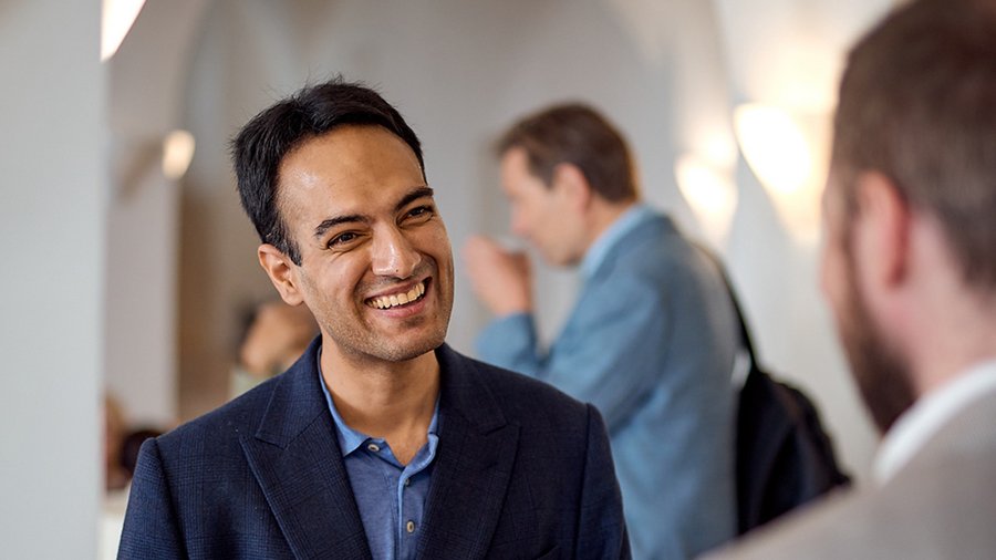 a man with dark hair stands smiling at another man who is out of focus