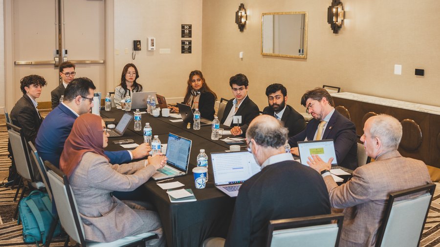 a group of people sit around a table in discussion while working on laptops