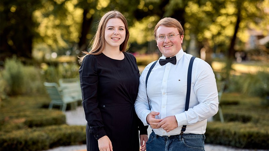 a woman with brown hair wearing a black dress stands next to a man wearing a white shirt. both are smiling at the camera with a garden in the background
