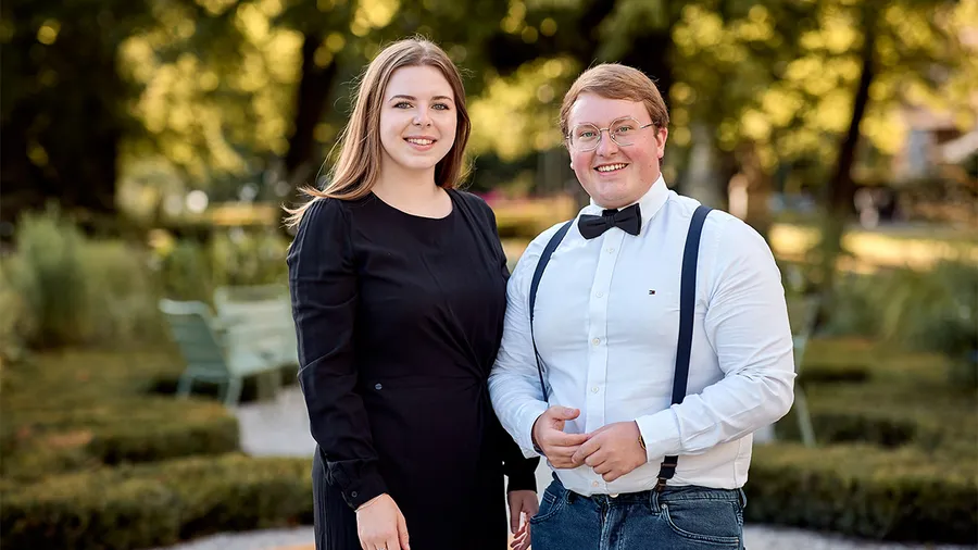 a woman with brown hair wearing a black dress stands next to a man wearing a white shirt. both are smiling at the camera with a garden in the background