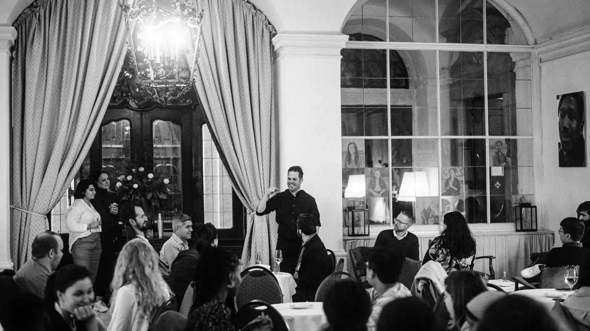 Black and White Photo of a Group of People sitting on Chairs in the Schloss and Listening to a Man Standing and Explaining