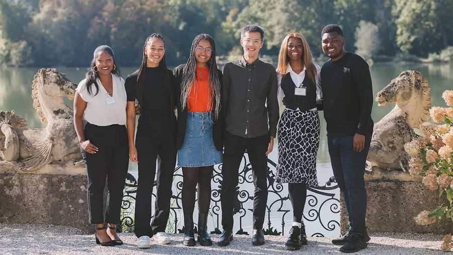 A group of six people stand in front of a lake and trees smiling at the camera.