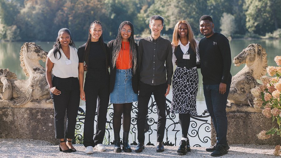 A group of six people stand in front of a lake and trees smiling at the camera.