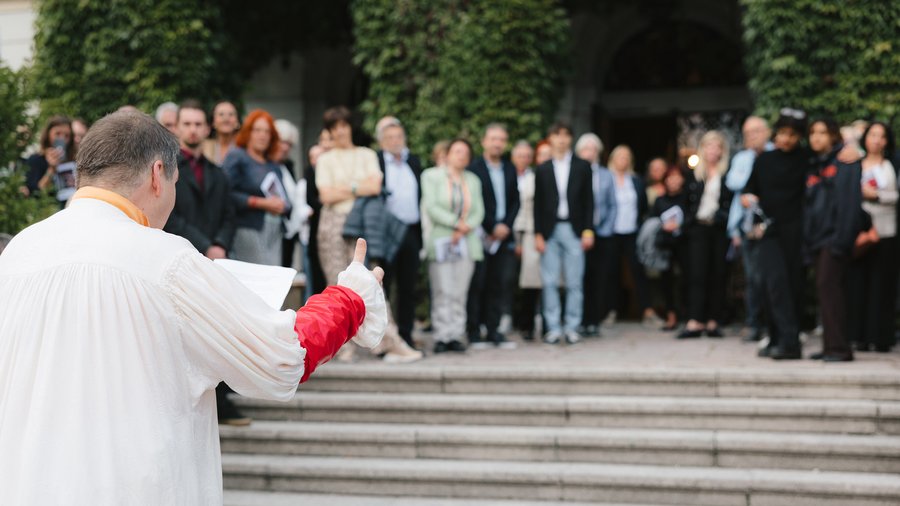 A man dressed in white reads from a paper in front of an onlooking crowd standing in front of Schloss Leopoldskron
