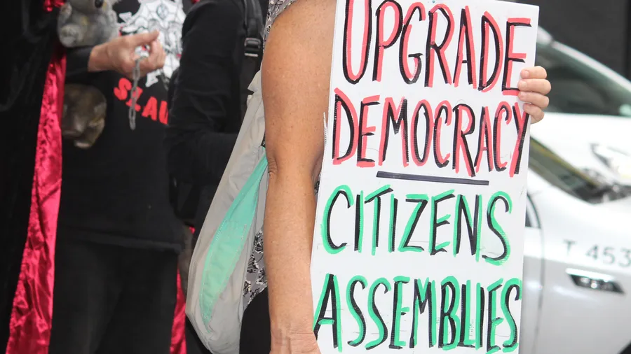 An image of a woman holding a sign reading "Upgrade Democracies: Citizens Assemblies"