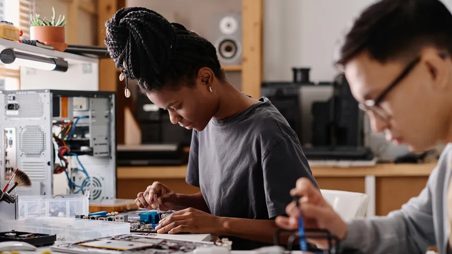 Two young adults work at a desk, surrounded by computer parts. A woman with dark braided hair repairs a circuit board with a small screwdriver. Another person works on equipment. The workshop setting is filled with tools, computer components, and bright indoor lighting.