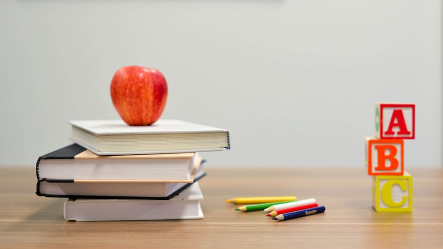 A red apple is balanced on top of some school books. Next to it on the right are some coloring pencils and A B C building blocks.