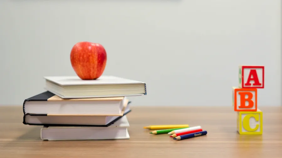 A red apple is balanced on top of some school books. Next to it on the right are some coloring pencils and A B C building blocks.