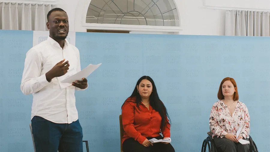 A man stands on the left holding a microphone and speaking with two women sitting next to him on the right
