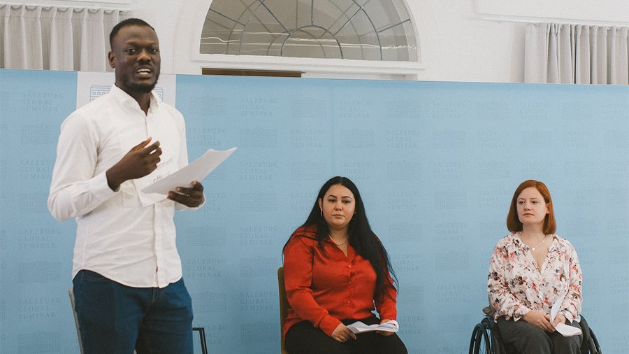 A man stands on the left holding a microphone and speaking with two women sitting next to him on the right