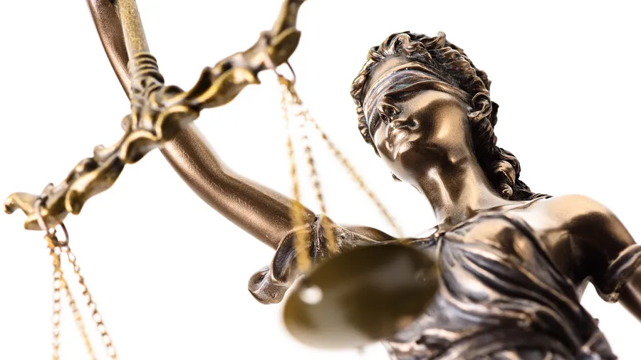 A low angle shot presents a bronze statue of Lady Justice holding scales. The statue is set against a bright white background. The statue appears to be a woman with curly hair and a blindfold, wearing draped clothing. The scales are suspended from a rod.