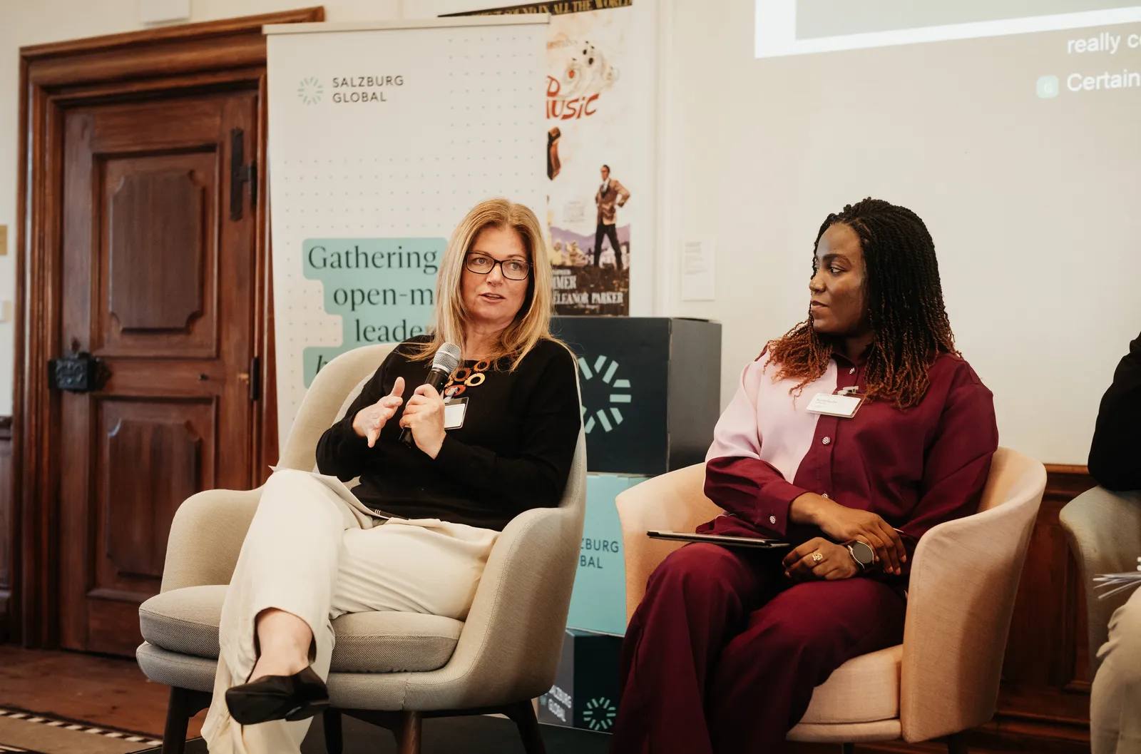 two women sit on stage while the one on the left speaks into a microphone