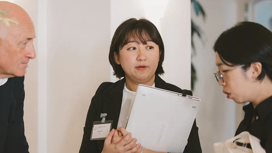 a woman holding a laptop stands in the middle of two other people during a conversation