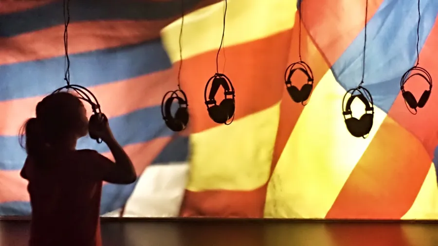 A child's silhouette is visible in the photo as they raise a pair of headphones to their head. Several more pairs of headphones are suspended above the child's head. In the background, a pattern of red, blue, and yellow stripes is displayed.