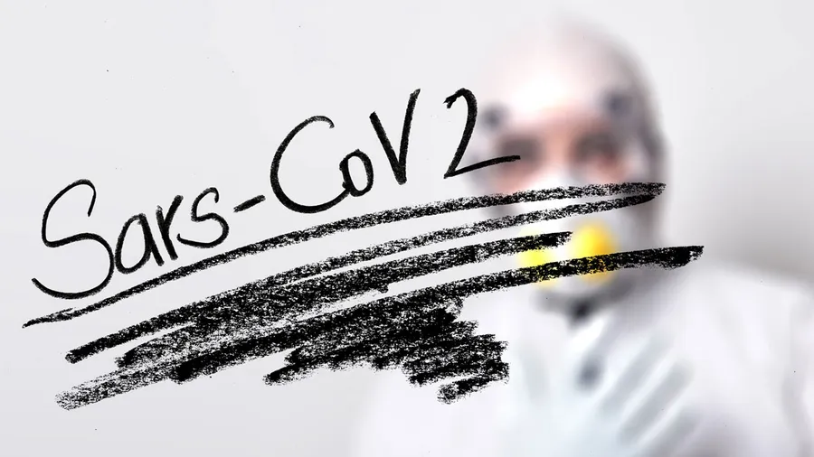In the forefront, Sars-Cov2 written in black ink on a perspex screen. In the background, a person wearing forensic clothing. 