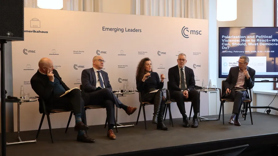 five individuals sitting on a stage, with a woman in the middle speaking into the microphone