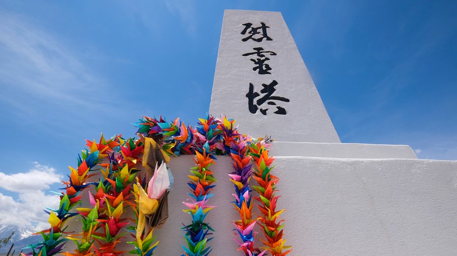 White memorial monument with Japanese inscriptions, decorated with colorful origami crane garlands under a bright blue sky