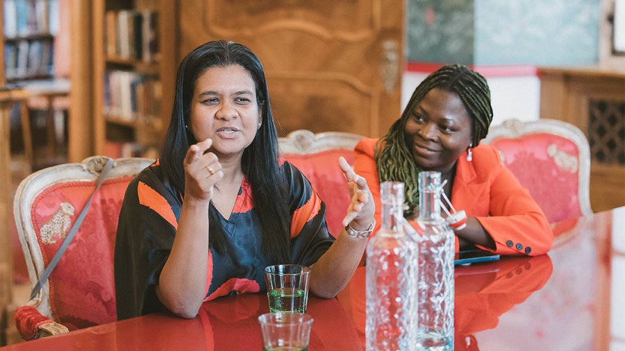 Two women sit at a red table, with the one on the left speaking and the one on the right listening. 