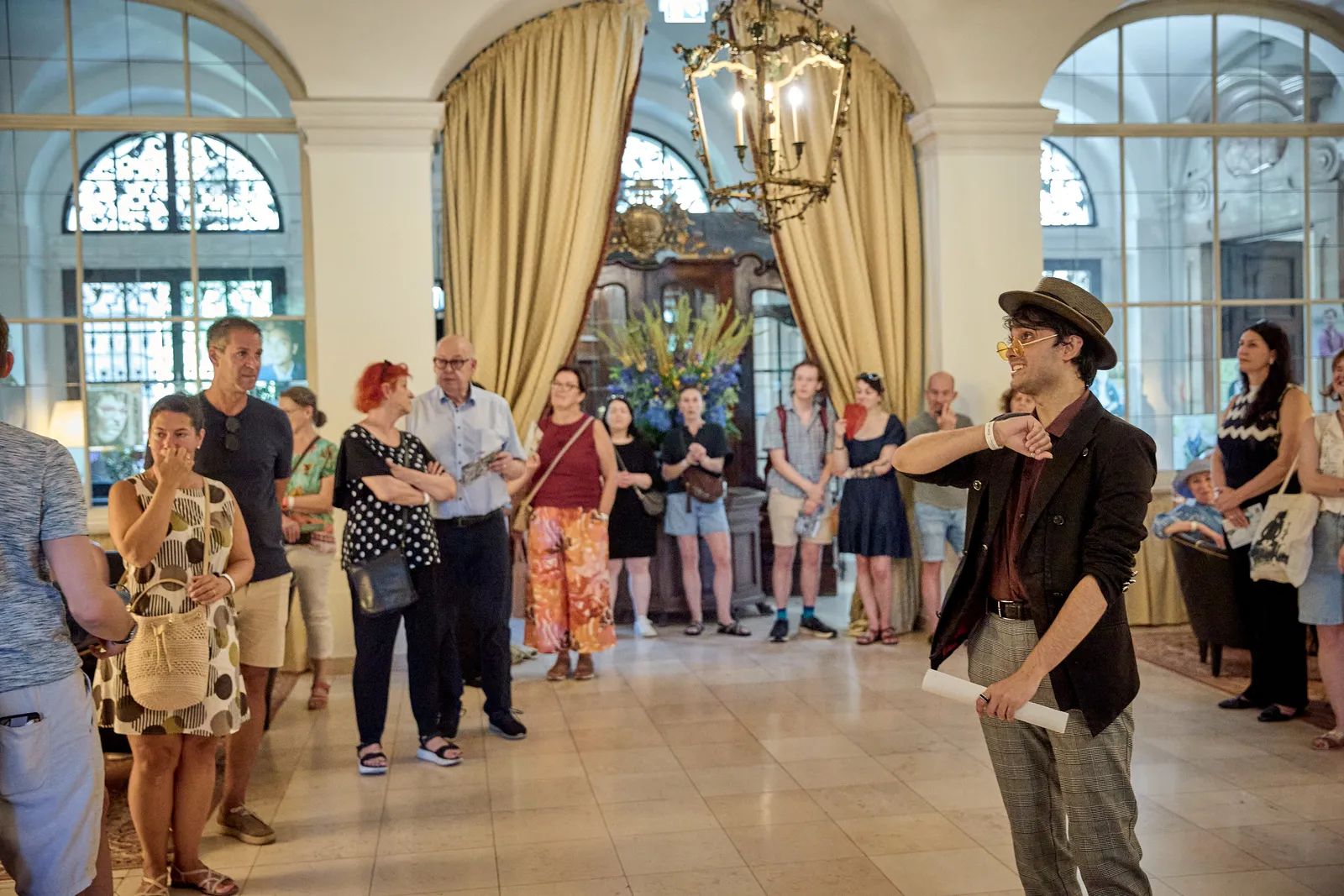 a man wearing a red jacket and hat speaks to a group, all of whom are standing in the Great Hall