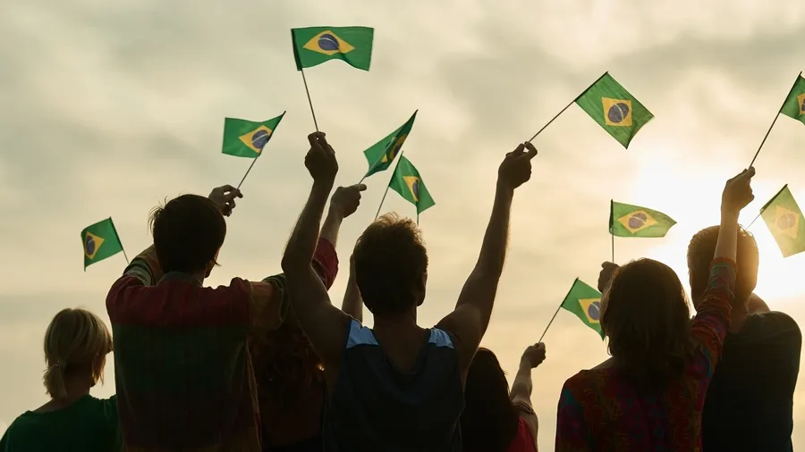 A large group of people are seen from behind, silhouetted against a bright sky at sunrise or sunset. The group members are raising their arms and waving flags. The photo evokes a feeling of unity and celebration.