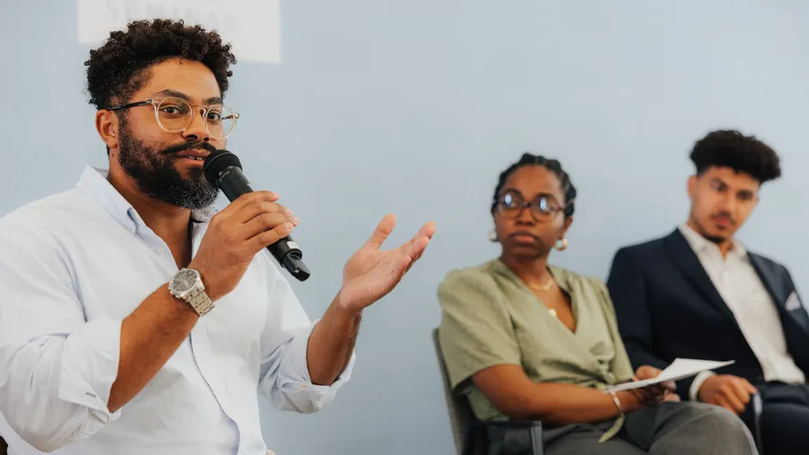 A man with dark hair, a beard, and glasses speaks into a microphone with two people listening attentively in the background.