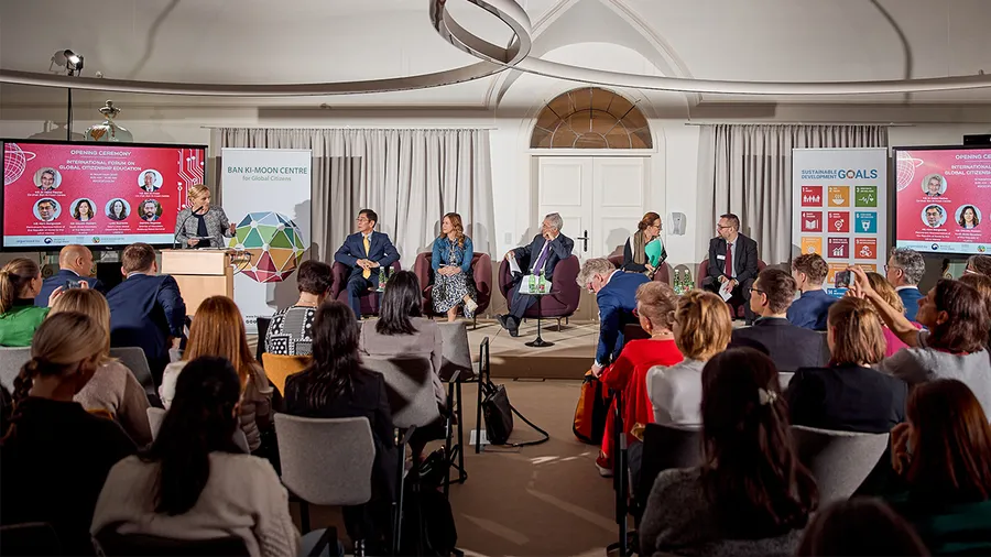 An audience sits in front of a panel of 5 speakers and a woman stands speaking at a podium on the left side