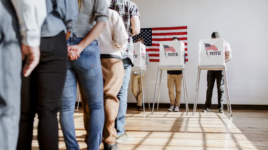 Several people are standing in line to vote indoors, the setting is bright and airy. The people are wearing various casual clothes, including jeans, t-shirts, and button-down shirts. Two voting booths are visible, each with a sign that reads "Vote" next to a small United States flag. Another larger United States flag is on the wall in the background. 