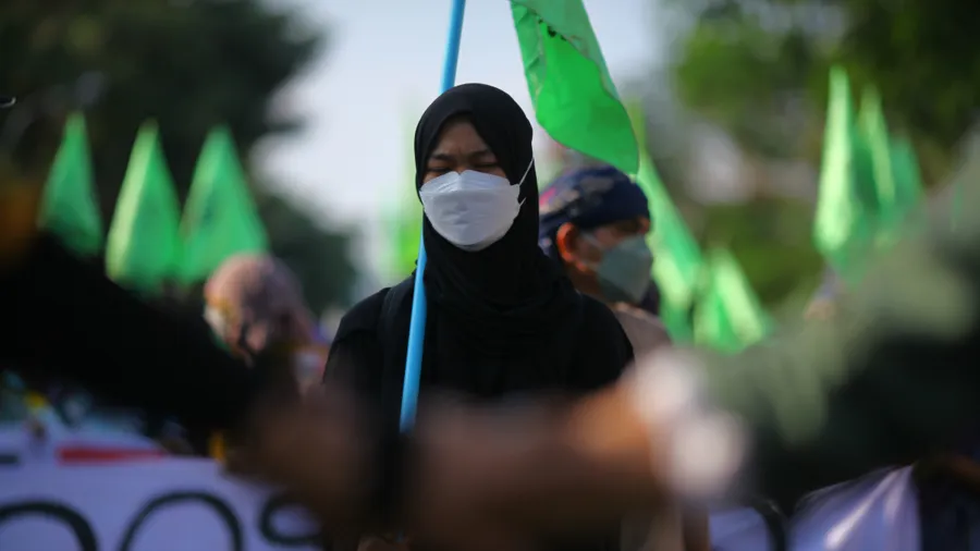 a woman wears all black with a white face mask and looks downward while protesting on the street