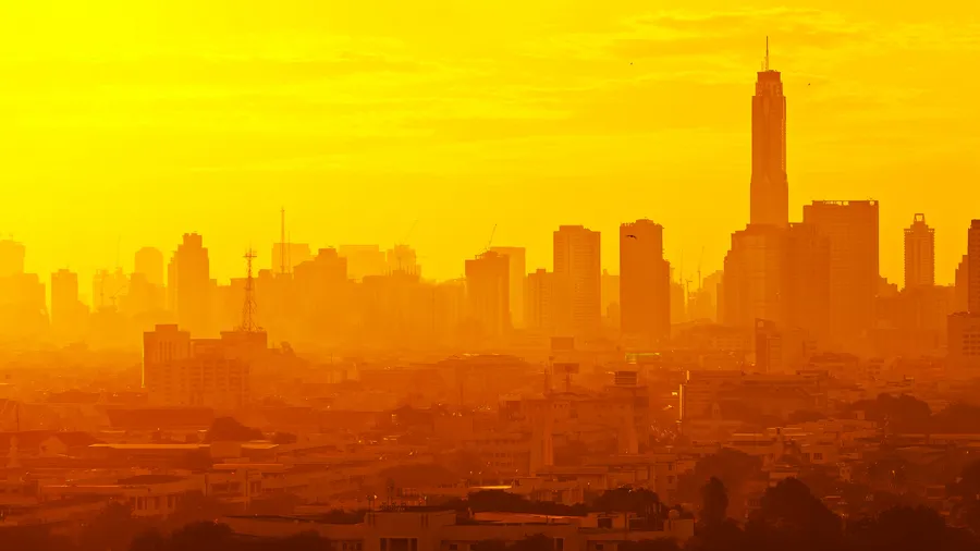 A panoramic vista reveals an urban city skyline bathed in the radiant light of sunrise or sunset. The warm, dreamy atmosphere is created by the sunlight, casting an orange glow over the buildings. Mist and shadows add depth to the cityscape.