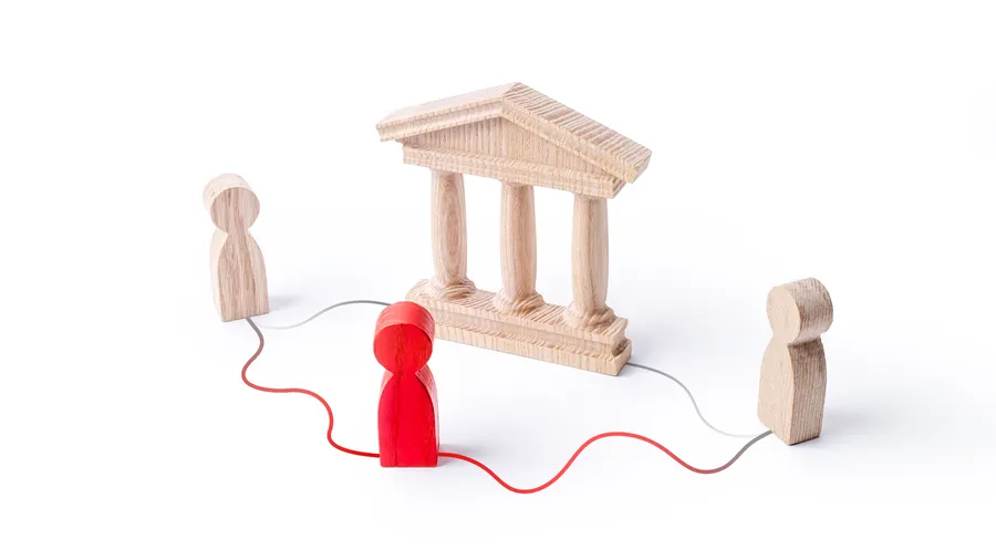 Three wooden figures stand on a white surface, connected by lines of red and gray, with a wooden model of a courthouse behind one.