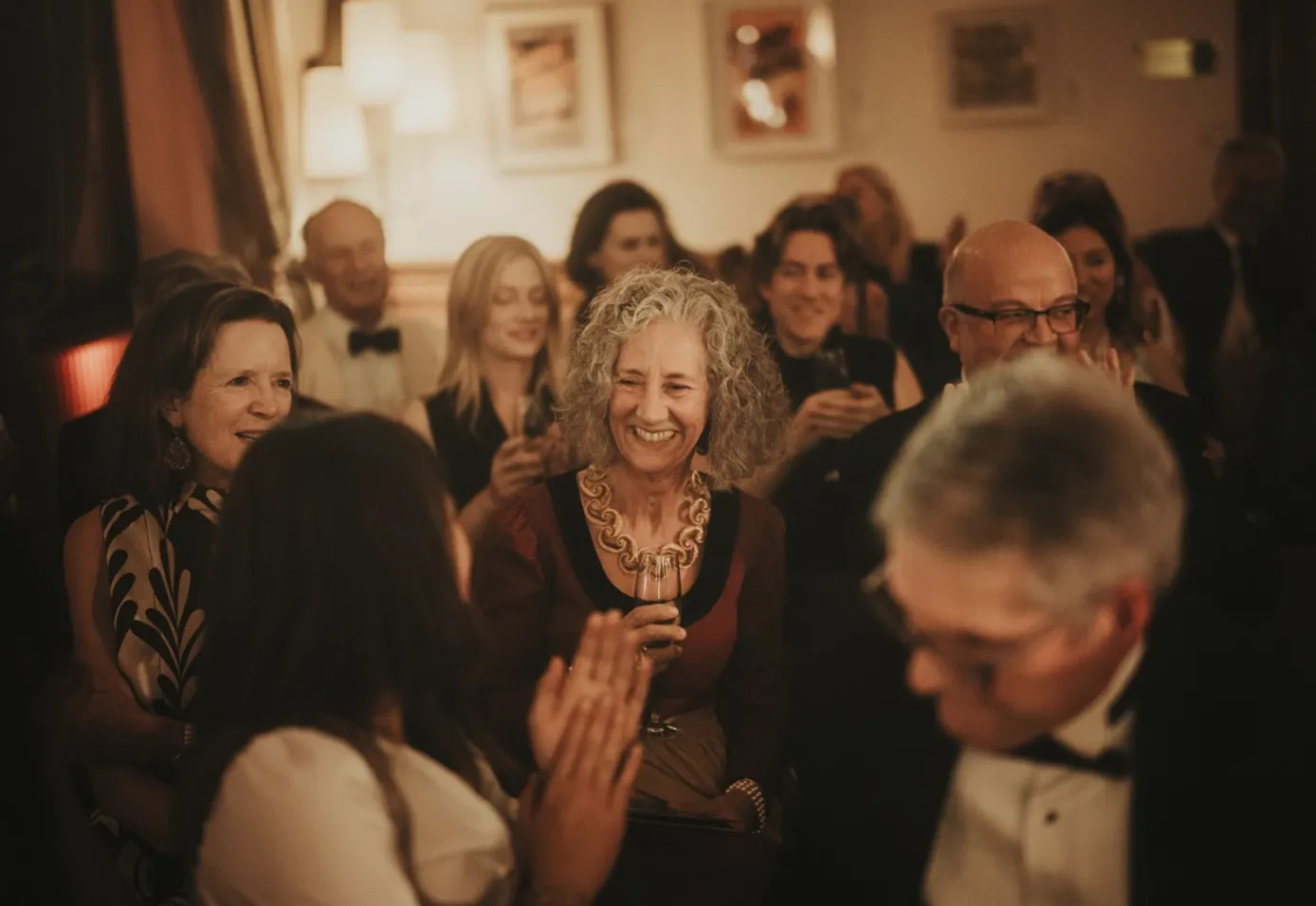 a woman with curly gray hair, smiling, holds a wine glass while those around her clap