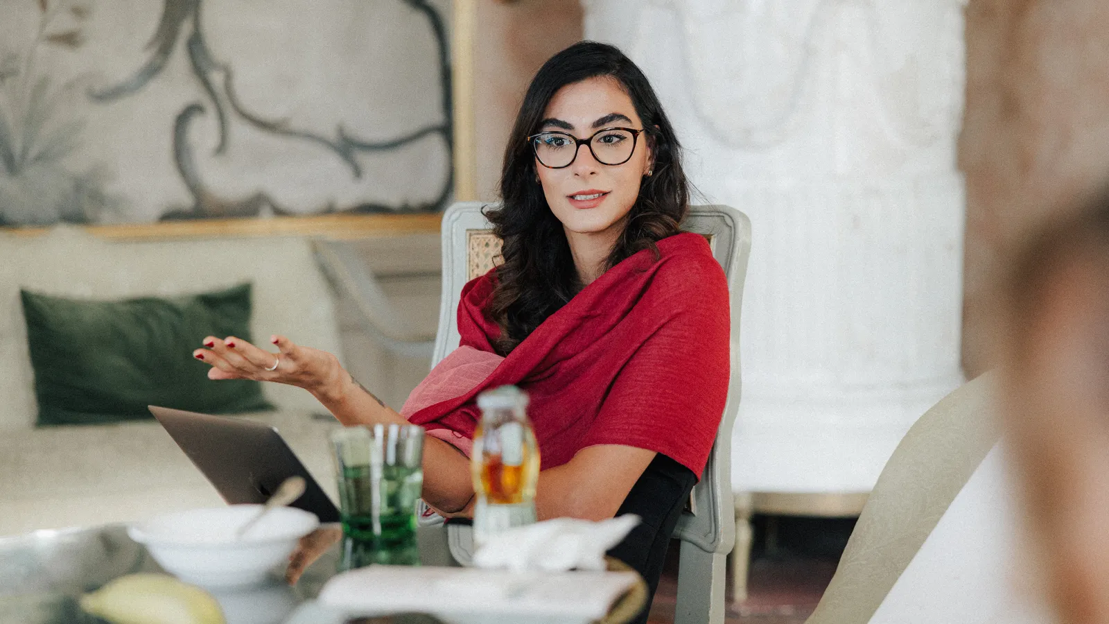 A woman with dark hair and glasses wearing a red scarf is speaking.