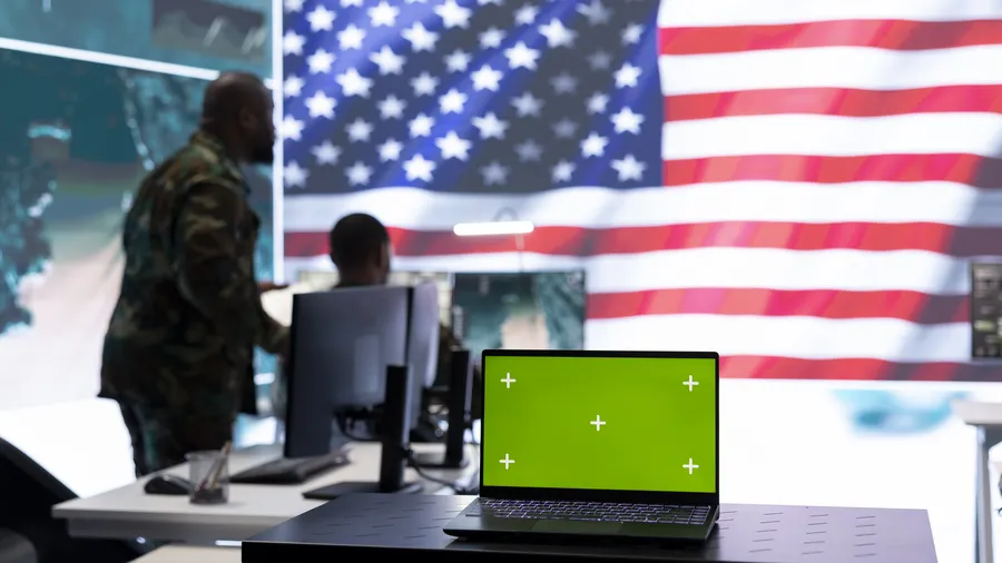 A modern office setting depicts two people working at computer stations and one person standing nearby. A laptop with a green screen sits on a desk in the foreground. Large monitors display a map and the American flag.