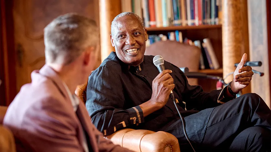 The image shows a smiling man holding a microphone while speaking and gesturing with one hand. He is dressed in a black shirt and sits in a cozy setting with a bookshelf in the background. Another person, slightly blurred, listens attentively to him.