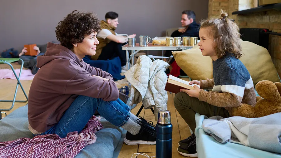 Young woman in casualwear listening to her little son with open book reading it and discussing with his mother in room for refugees