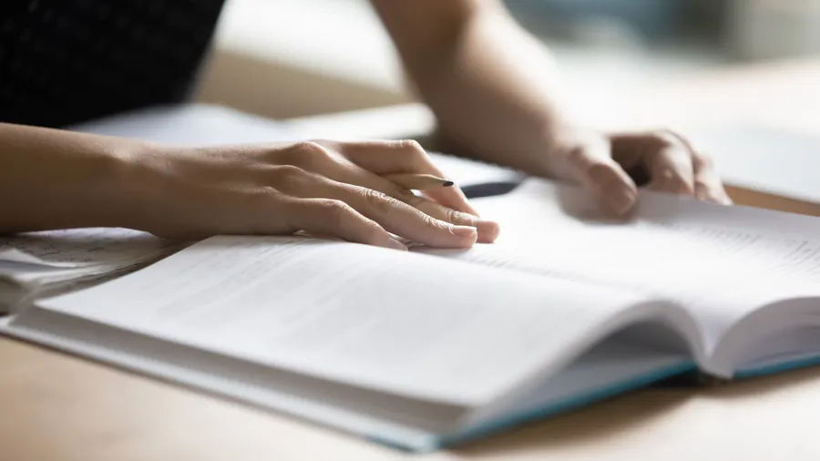 Close-up photo of a person's hands holding a pen and resting on an open textbook.